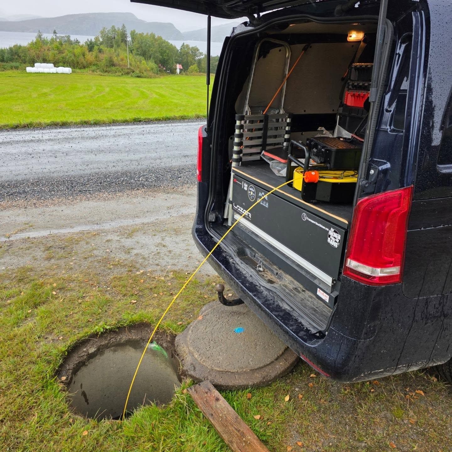 Åpen bakluke på servicebilen med utstyret om bord, med grasbakke og vann bak